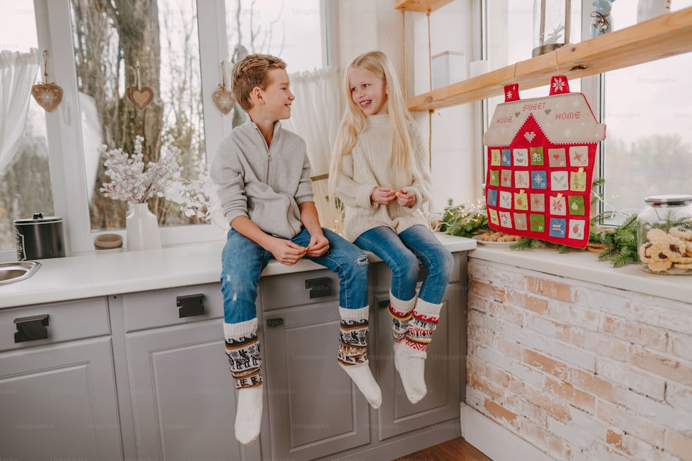 家の形をしたクリスマスの手作りアドベントカレンダーの近くのキッチンに座って楽しんでいる幸せな男の子と女の子。