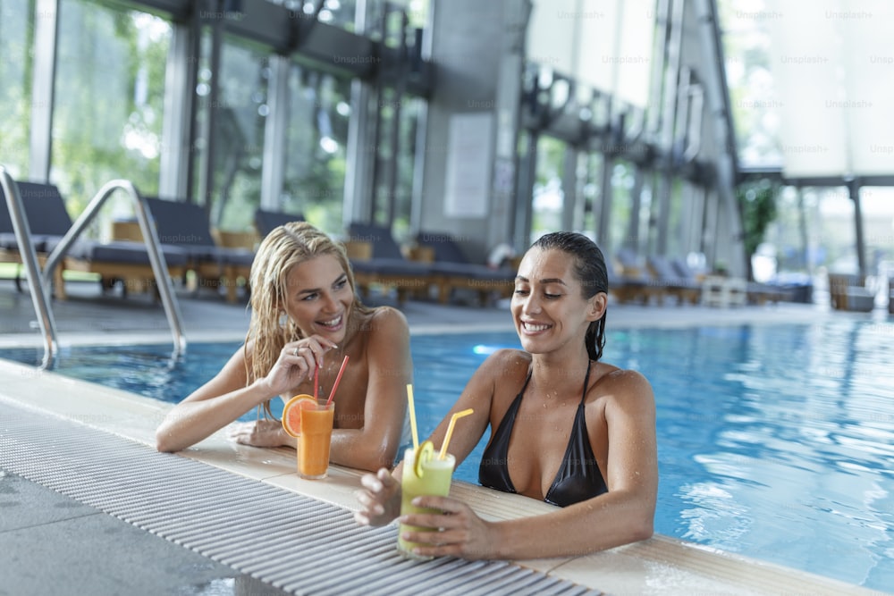 女性の友人は、プールバーでカクテルモヒートを飲み、熱帯の島のビーチ近くのビキニの高級ホテルを着てリラックスします。プールで楽しんだり、カクテルを飲んだり、笑顔を浮かべている美しい若い女性。