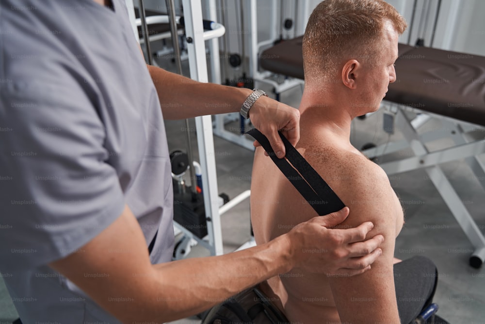 男性の患者の肩にテープを貼るプロの理学療法士。背中の痛みのキネシオテーピング