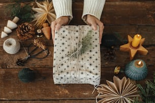 Mains tenant un cadeau de Noël élégant enveloppé dans du papier sur une table en bois rustique avec des décorations de Noël festives, vue de dessus. Cadeau de Noël et bougie, fil, ciseaux, pomme de pin, branches de sapin, étoile