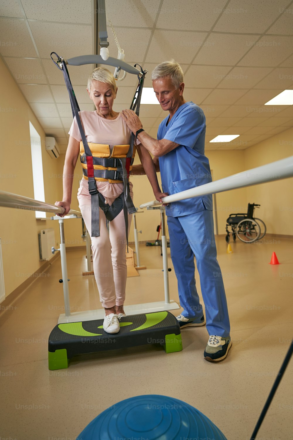 Stroke female patient using walking harness system and assisted by healthcare worker while exercising on step platform in rehabilitation clinic