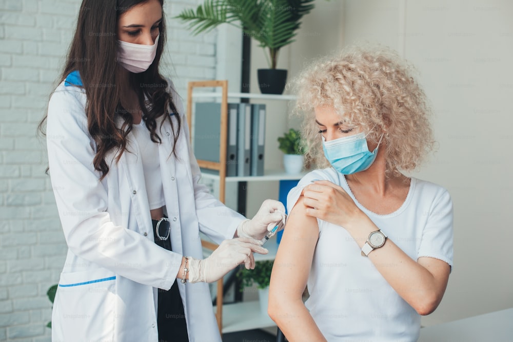 Side view photo of a blonde curly woman having a corona virus vaccine at the medical center from a medical sister with mask