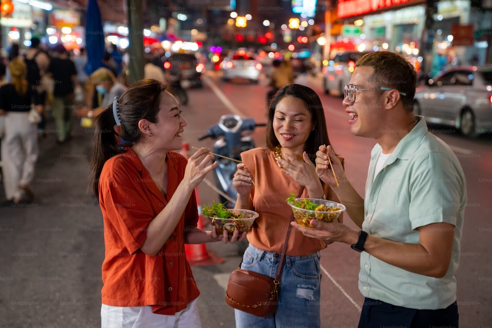 Grupo 4K de mulheres asiáticas e pessoas LGBTQ amigos turistas gostam de comer tradicional comida de rua churrasco frutos do mar lula grelhada com molho picante juntos no mercado noturno de rua da cidade da China em Bangkok, Tailândia