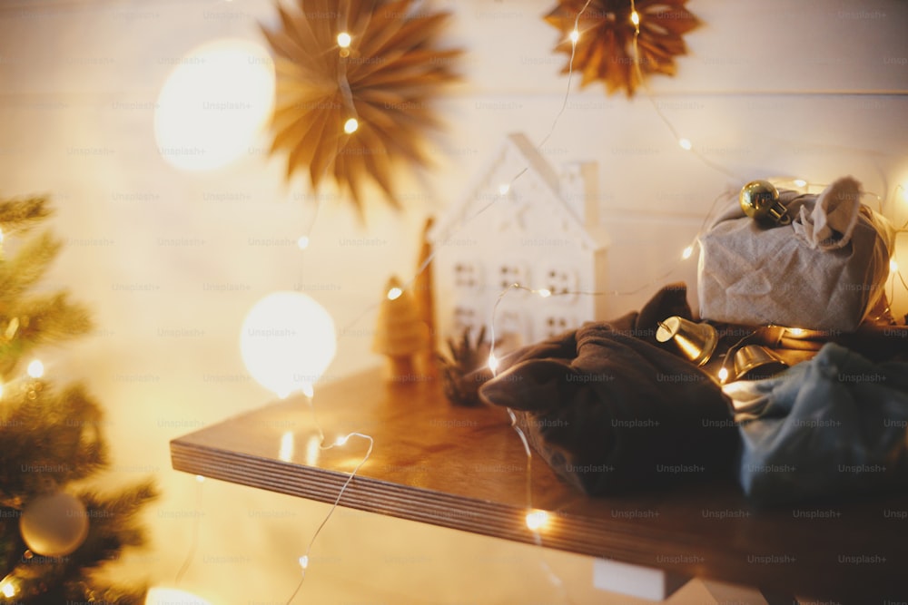 Stylish zero waste christmas gifts on wooden shelf on background of little house, trees, paper stars and christmas golden lights bokeh. Festive decorated scandinavian room. Atmospheric eve