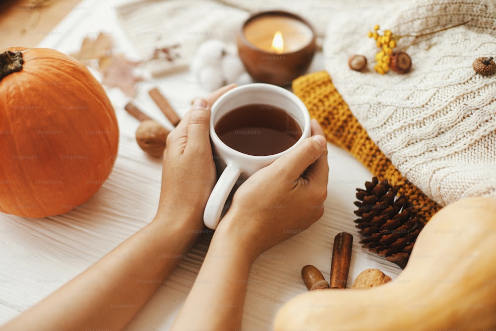 Hands holding warm cup of tea on background of autumn leaves, pumpkin, cozy sweaters, burning candle on white wood. Hello autumn and Happy Thanksgiving.