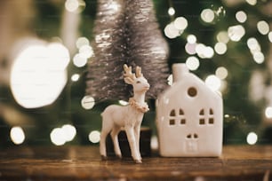 Merry christmas. Stylish christmas tree, reindeer toy and house on rustic wood on background of christmas lights. Beautiful little christmas scene. Magic christmas time. Space for text