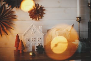 Stylish little christmas trees, house, plastic free gifts on background of white wooden wall with paper stars and golden lights bokeh. Space for text. Festive decorated scandinavian room