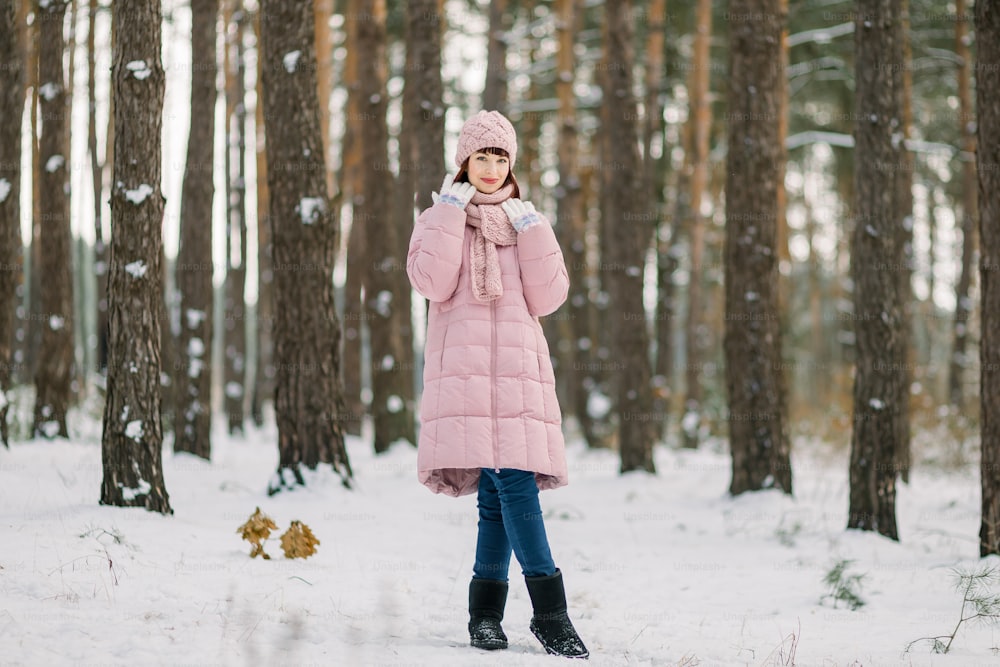 Graziosa donna di 30 anni in elegante cappotto rosa, berretto e sciarpa, che si gode una passeggiata nella bellissima foresta invernale innevata. Ritratto invernale all'aperto stagionale di felice signora caucasica