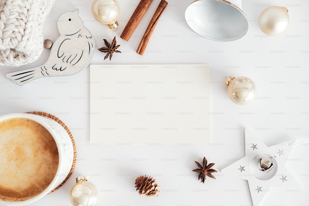 Mockup di biglietto d'auguri vuoto, bastoncini di cannella, decorazioni natalizie, tazza di cioccolata calda su tavolo da scrivania bianco. Hygge, casa accogliente, concetto di stile nordico.