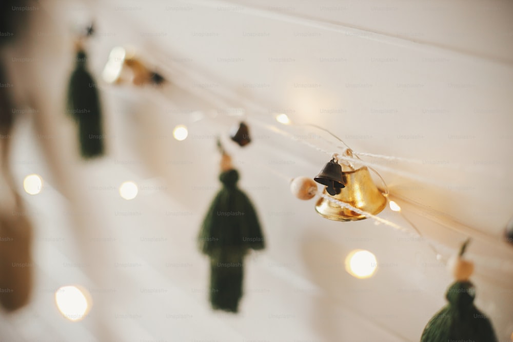 Stilvolle Weihnachtsgirlande hautnah mit Glocken und Bäumen an weißer Holzwand mit Weihnachtsbeleuchtung. Festlich dekoriertes stimmungsvolles skandinavisches Zimmer. Modernes Boho handgefertigtes Dekor. Platz für Text