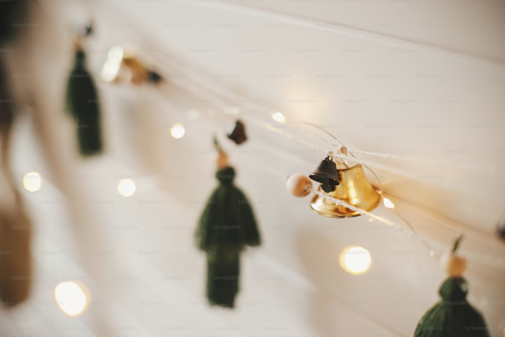 Stylish christmas garland close up with bells and trees on white wooden wall with christmas lights. Festive decorated atmospheric scandinavian room. Modern boho handmade decor. Space for text