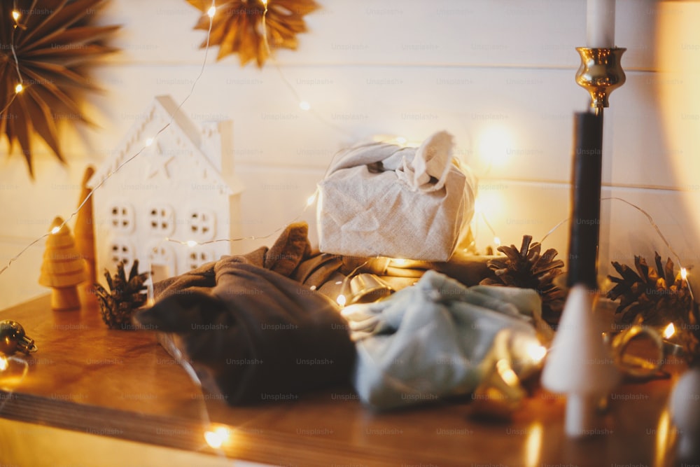 Stylish zero waste christmas gifts on wooden shelf on background of little house, trees, paper stars and christmas golden lights bokeh. Festive decorated scandinavian room. Atmospheric eve