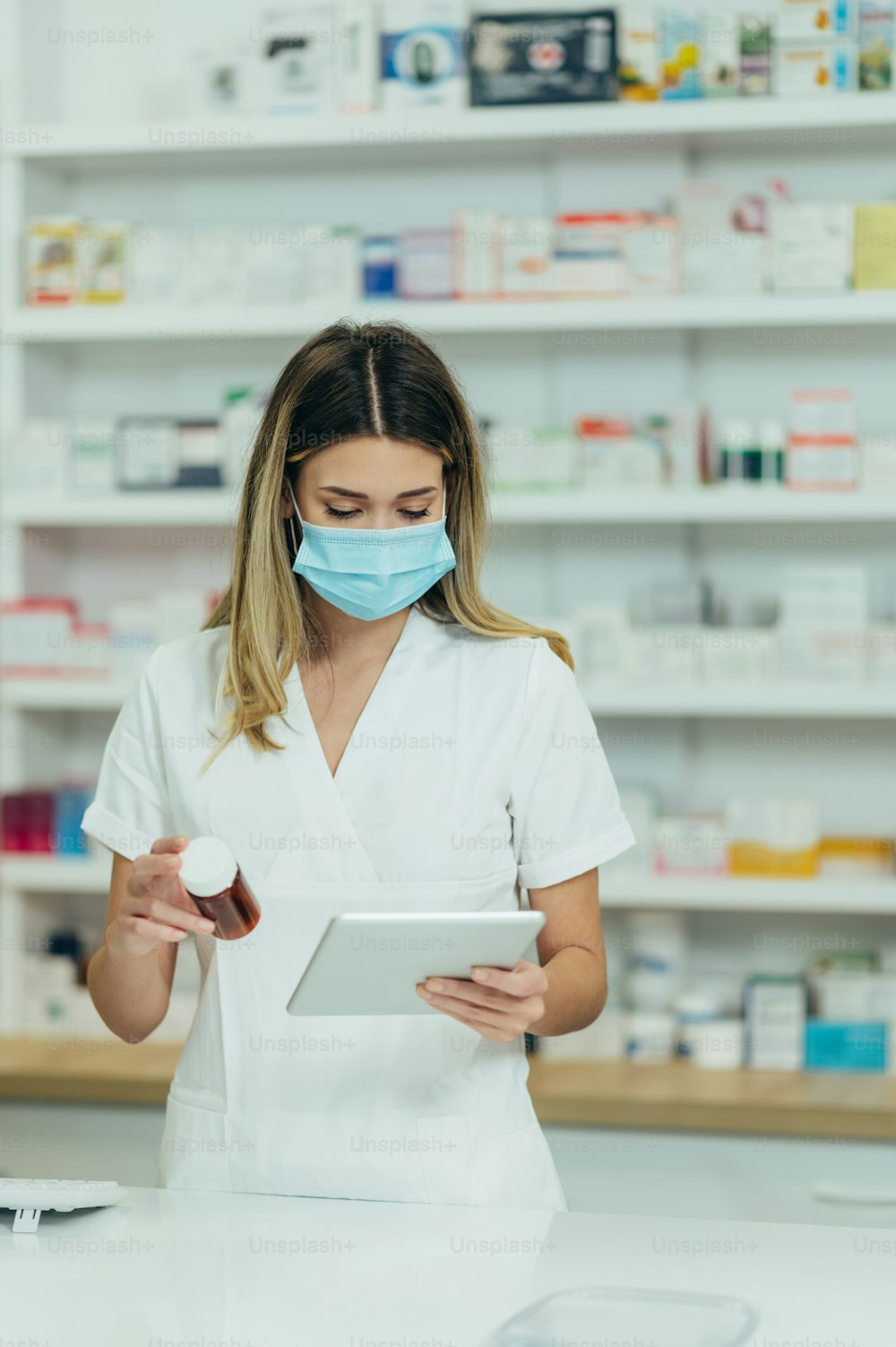 Hermosa farmacéutica con máscara protectora en la cara sosteniendo medicamentos y usando una tableta digital mientras trabaja en una farmacia