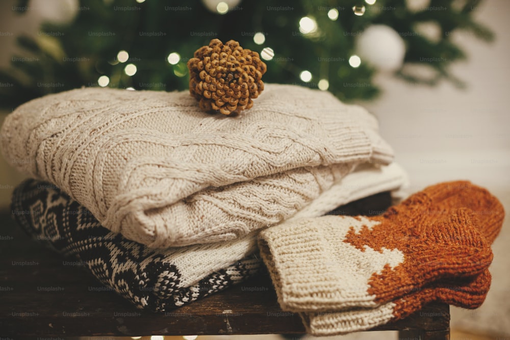 Cozy knitted sweaters and wool socks on rustic wood with pine cone on background of christmas tree lights. Pile of stylish winter cloth in festive decorated scandinavian room. Winter hygge time