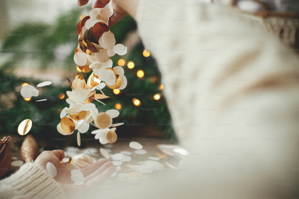 Manos lanzando confeti dorado sobre fondo de luces navideñas, decoraciones, adornos y ramas de pino sobre madera rústica. Espacio para el texto. Felices Fiestas. Fiesta de año nuevo y navidad. Ambiente festivo