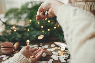 Hands in sweater holding golden confetti on background of christmas lights, decorations, baubles and pine branches on rustic wood. Festive mood. Space for text. Happy holidays. Xmas party