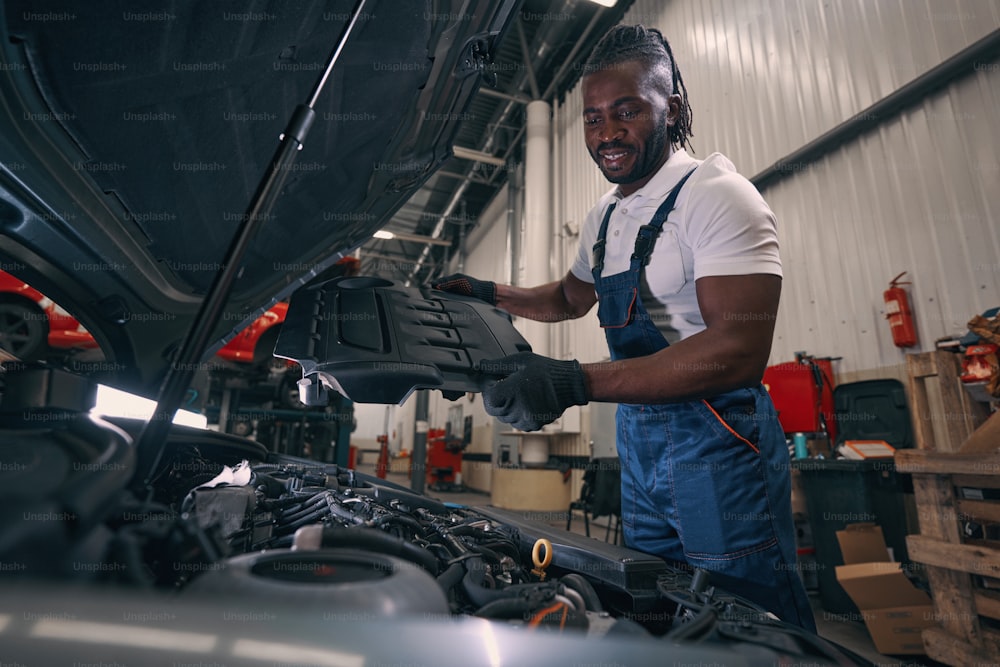 Pleased automotive technician removing protective cover from car engine and looking at its parts