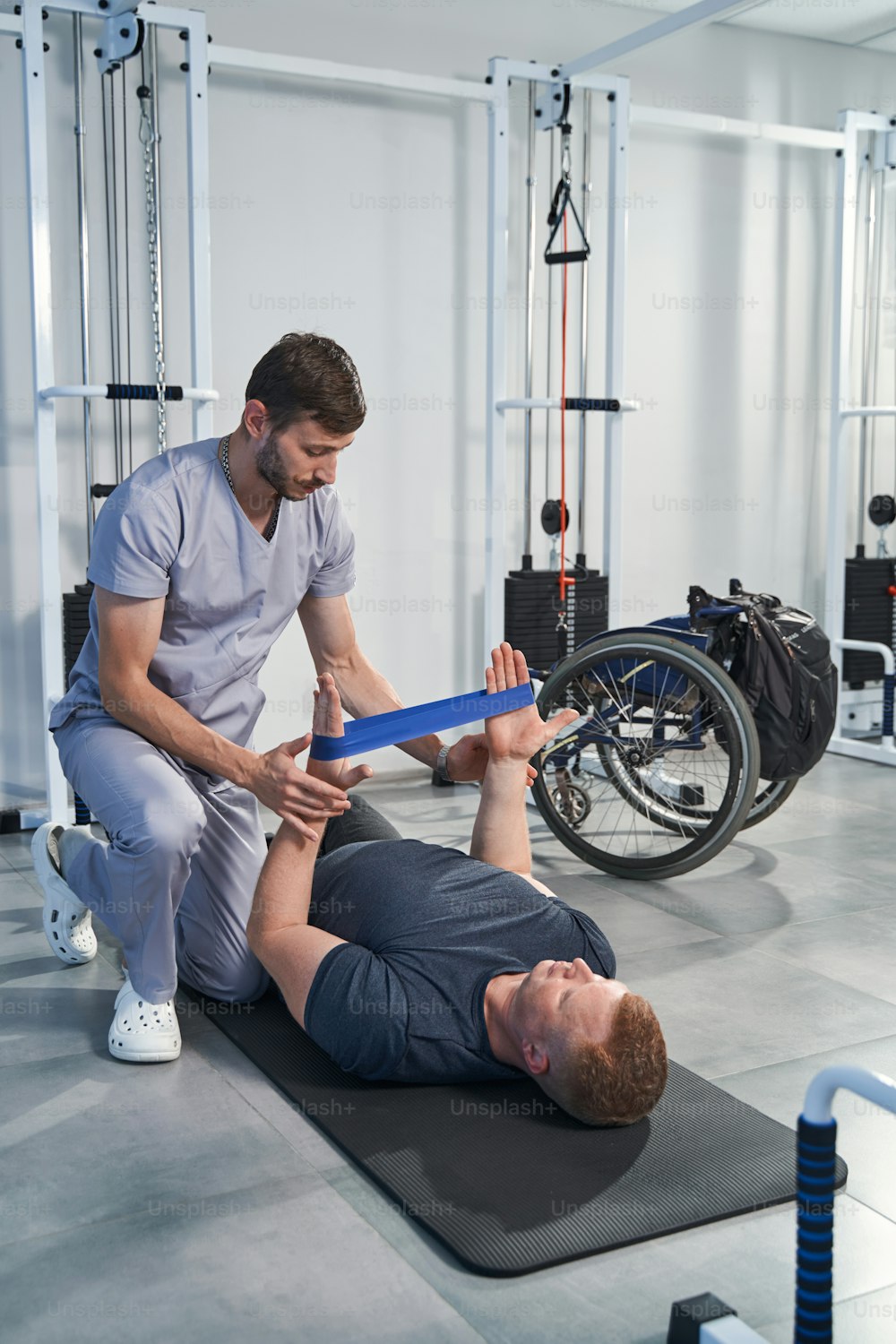 Male physiotherapist holds elastic band while man lying on mat for strength rehabilitation exercise
