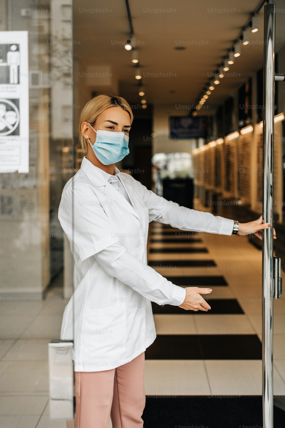 Giovane e attraente optometrista femminile con maschera protettiva per il viso in piedi alle porte aperte del negozio di ottica e guardando fuori. È sicura di sé e seria. Covid-19 aperto per il concetto di business.