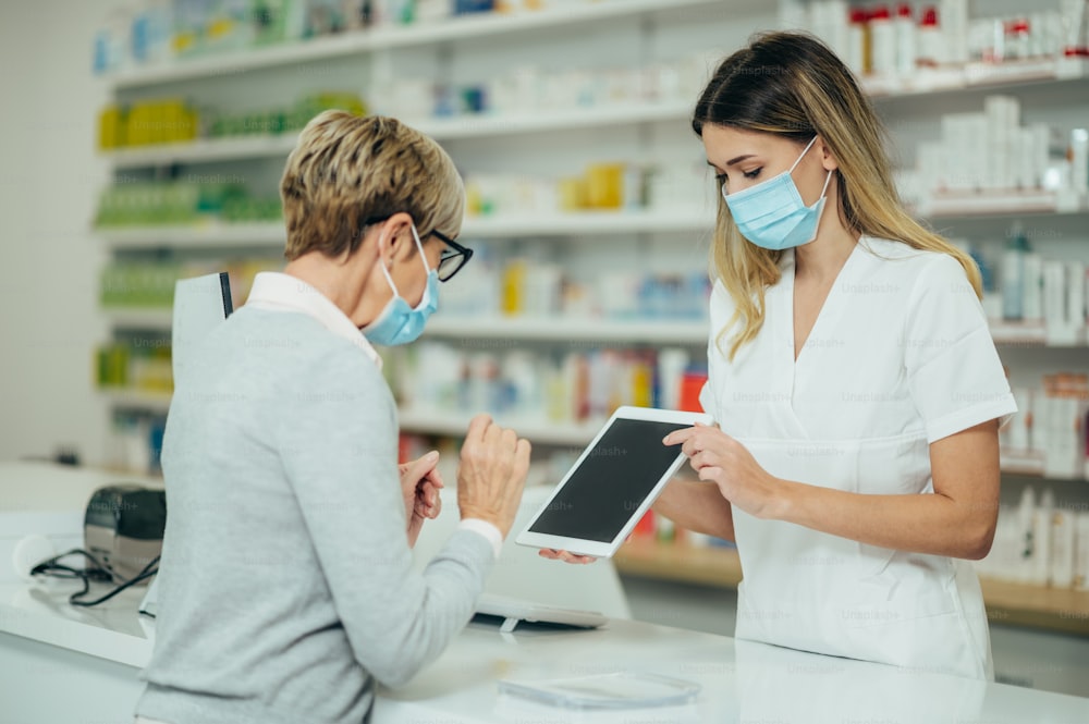 Farmacéutica con mascarilla protectora y atendiendo a un paciente cliente en una farmacia mientras muestra su tableta digital