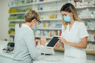 Farmacéutica con mascarilla protectora y atendiendo a un paciente cliente en una farmacia mientras muestra su tableta digital