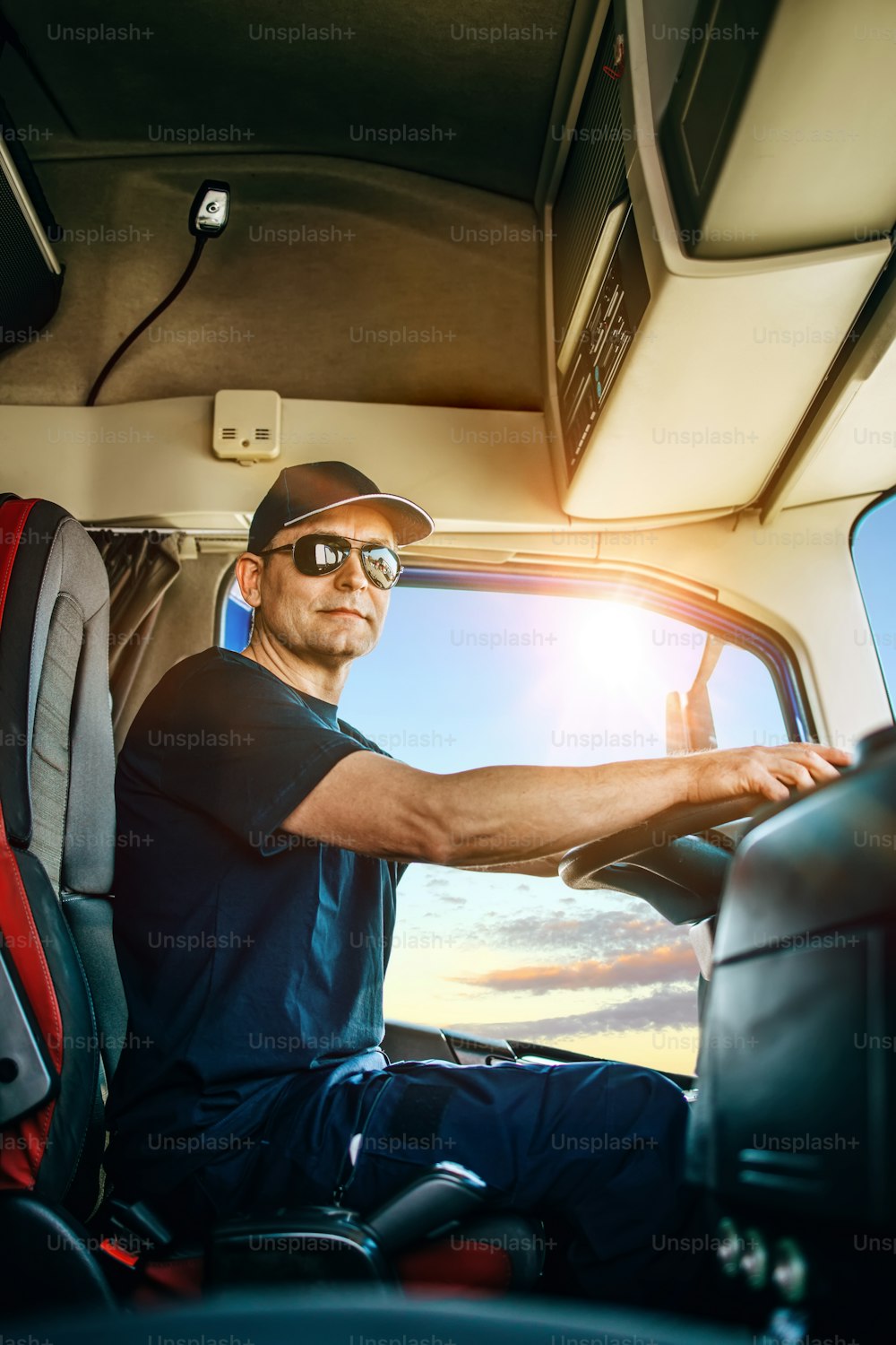 Handsome experienced male truck driver with a hat sitting and driving his truck. Professional transportation and truck drivers concept.