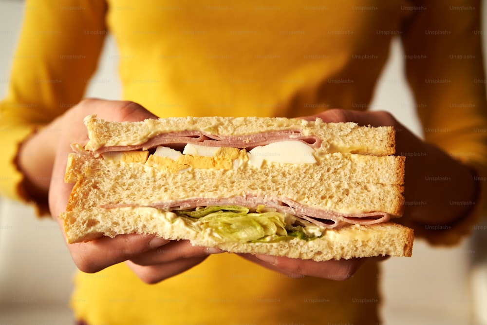 Sándwich con jamón, huevos y lechuga sostenido por las manos de una adolescente con top amarillo