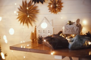 Stylish christmas gifts on wooden shelf on background of little house, trees, paper stars and christmas golden lights bokeh. Atmospheric festive scandinavian room. Eco friendly presents in eve