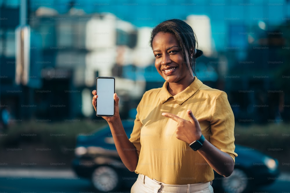 Schöne junge afroamerikanische Frau, die ein Smartphone zeigt, während sie auf der Straße der Stadt steht
