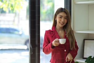 Gorgeous asian young businesswoman in red suit sipping hot coffee at her modern office.