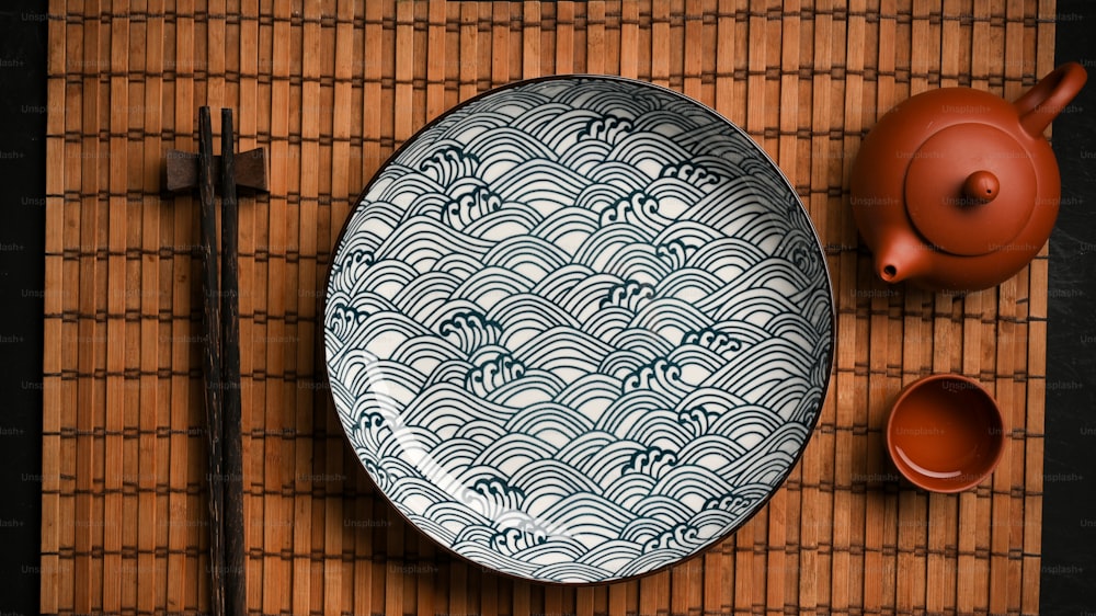 Asiatischer Porzellan-Keramikteller mit Essstäbchen und Teeset auf Bambus-Tischset. Draufsicht, flache Verlegung