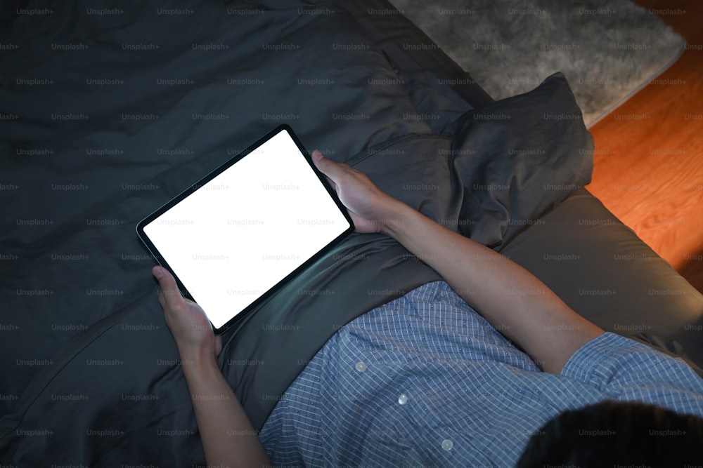 Young man lying under blanket and using digital tablet at night.