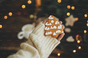 Hand in cozy sweater holding christmas tree gingerbread cookie on rustic moody background with warm lights. Winter hygge. Merry Christmas and Seasons greeting. Moody image