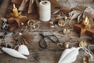 Stylish Christmas bells, paper angel and stars, ornaments, candle, thread and scissors on rustic wooden background. Making festive scandinavian garland, atmospheric christmas decor.