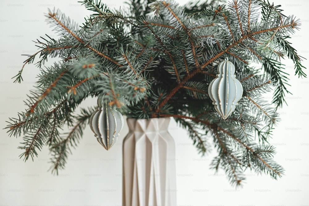 Stylish christmas ornament on fir branch in modern vase close up. Modern wooden bauble on branch on white background. Festive simple and creative decor in scandinavian room. Merry Christmas!