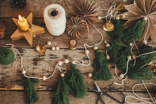 素朴な木製の背景にスタイリッシュなクリスマスモミの木の花輪、鐘、紙の星、ろうそく、糸、はさみ。フラットレイ。お祝いの手作りの休日の装飾を作ります。雰囲気のあるクリスマスタイム
