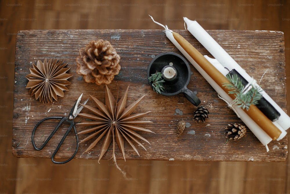 Stylish Christmas candles, paper stars, pine cones and fir branch, scissors on rustic wooden background. Christmas flat lay. Festive decor. Atmospheric Scandinavian hygge. Holiday advent