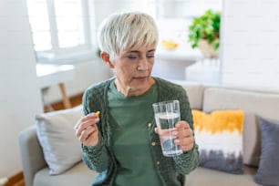 Concept of suffering from terrible pain. Mature, sad, upset, unhappy, troubled, weak woman with gray hair, wearing casual clothes is sitting on a sofa and taking a pill