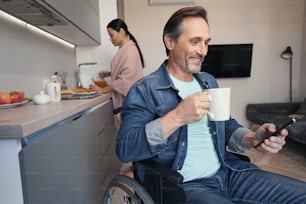 Happy mature male with disability is chatting on cell phone and drinking coffee while woman is tyding up