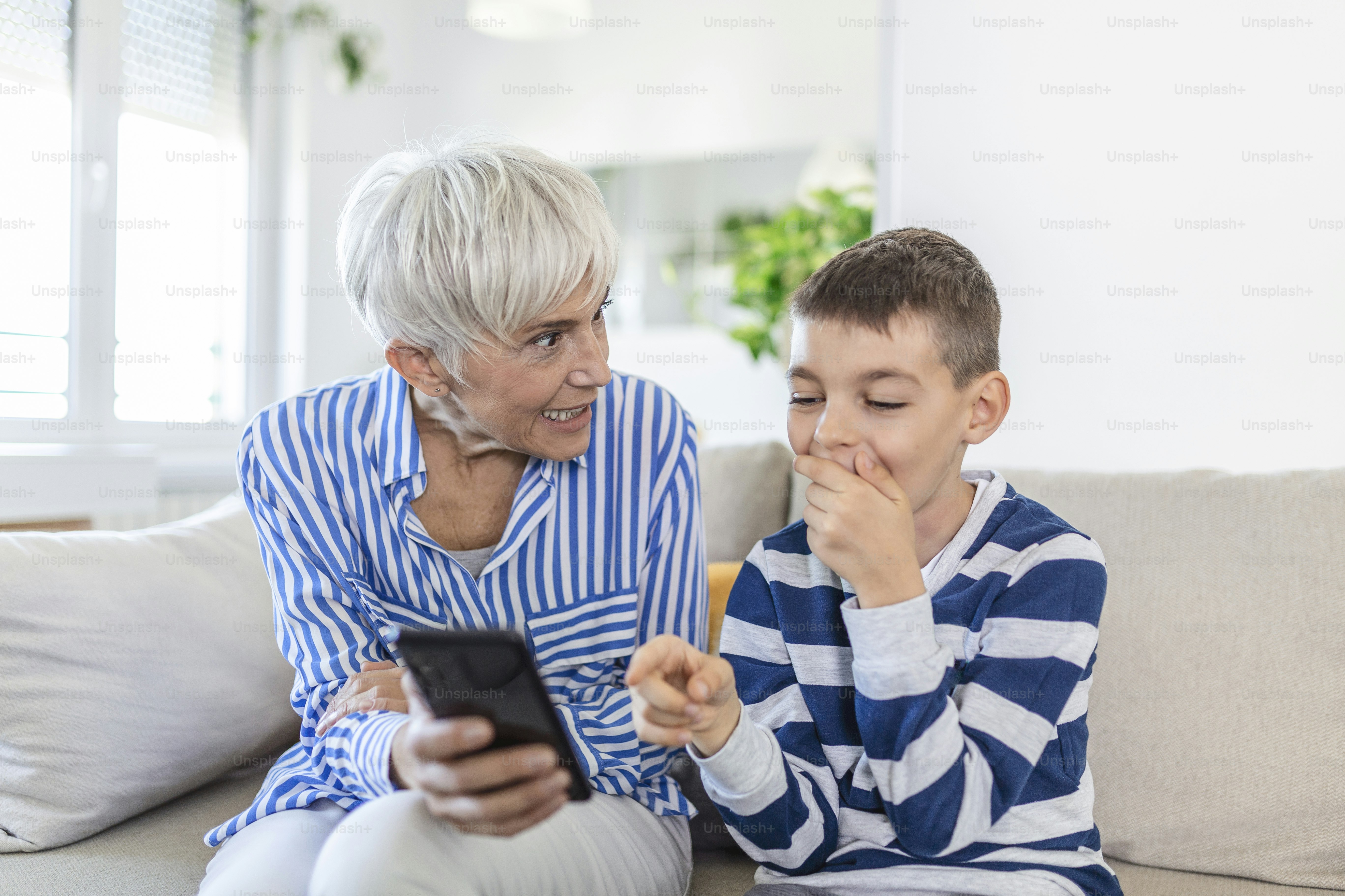 Happy grandmother and grandson using phone together, sitting on cozy sofa at home, browsing mobile device apps, grandma with grandchild playing game, looking at screen, having fun