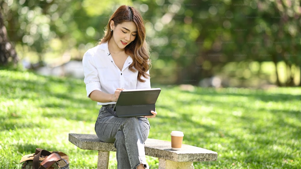 Fêmea encantadora sentada no banco com tablet do computador no parque público.