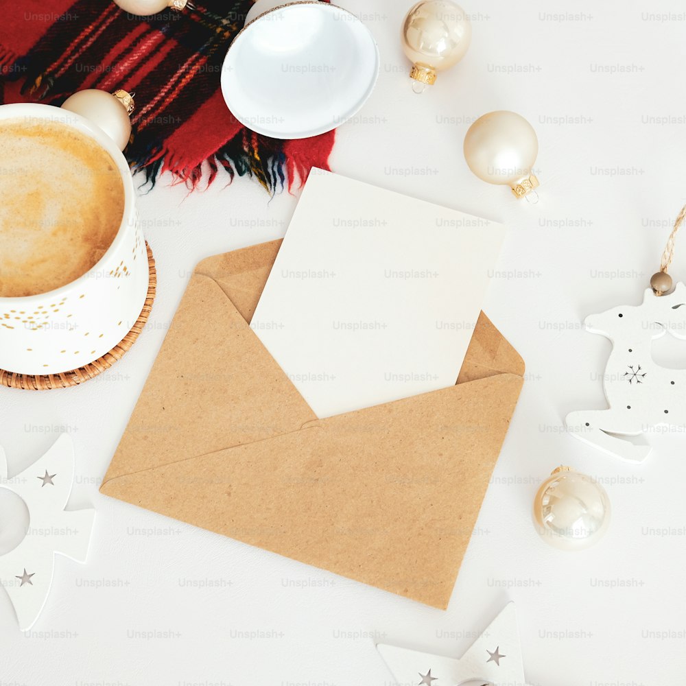 빈 인사말 카드, 커피 한 잔, 흰색 테이블에 크리스마스 장식이 있는 봉투. 메리 크리스마스와 새해 복 많이 받으세요 개념.