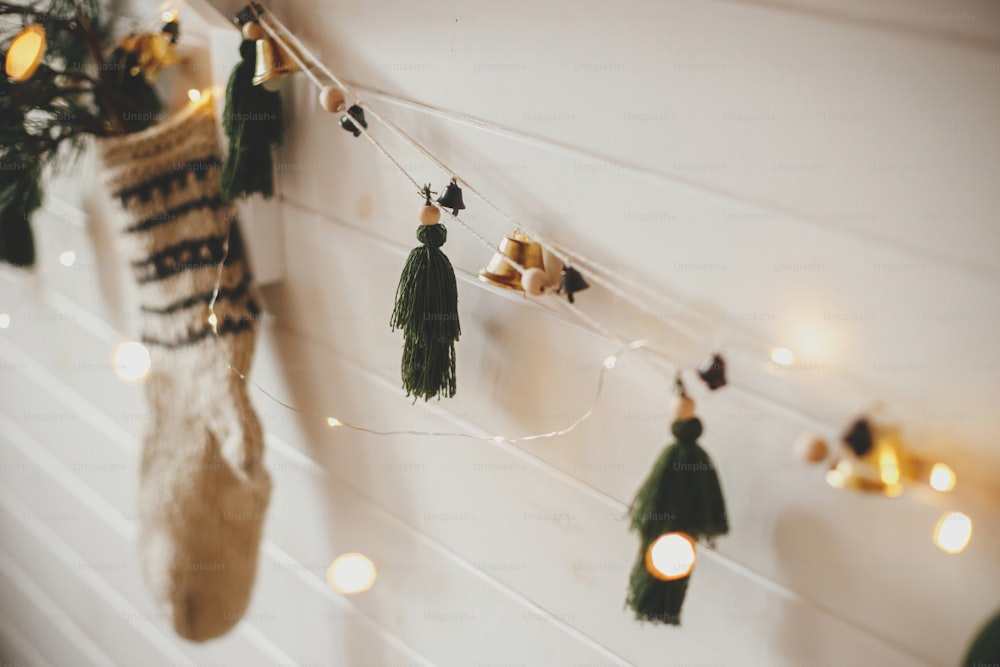 Stylish christmas garland with bells, rustic stocking with fir branch and christmas lights on wooden wall. Festive decorated atmospheric scandinavian room. Modern handmade decor. Space for text