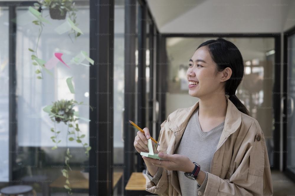 Smiling creative woman reading sticky notes on glass wall in modern workplace.