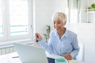 Focused old woman with white hair at home using laptop. Senior stylish entrepreneur working on computer at home. Woman analyzing and managing domestic bills and home finance