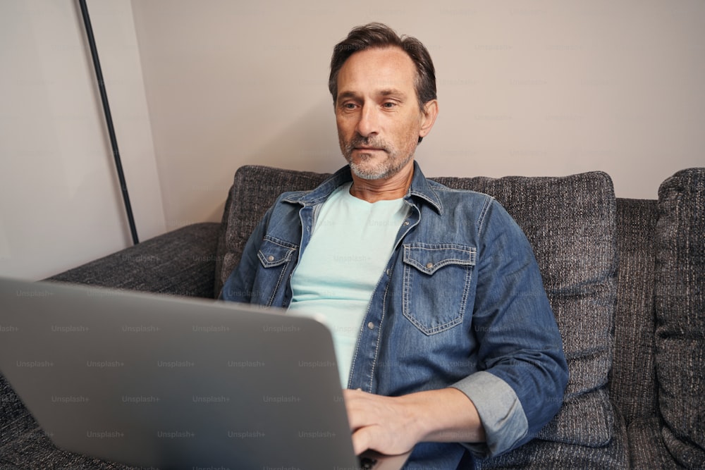 Un homme caucasien calme est assis sur le canapé et utilise un ordinateur portable tout en passant la journée seul à l’intérieur