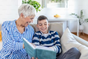 Cropped photo of happy gray-haired woman looking at book with her grandson indoors. They are smiling during their conversation. Visiting grandparents concept
