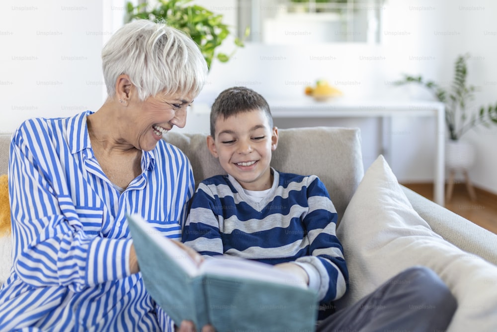 Beschnittenes Foto einer glücklichen grauhaarigen Frau, die mit ihrem Enkel drinnen ein Buch betrachtet. Sie lächeln während ihres Gesprächs. Großelternbesuch Konzept