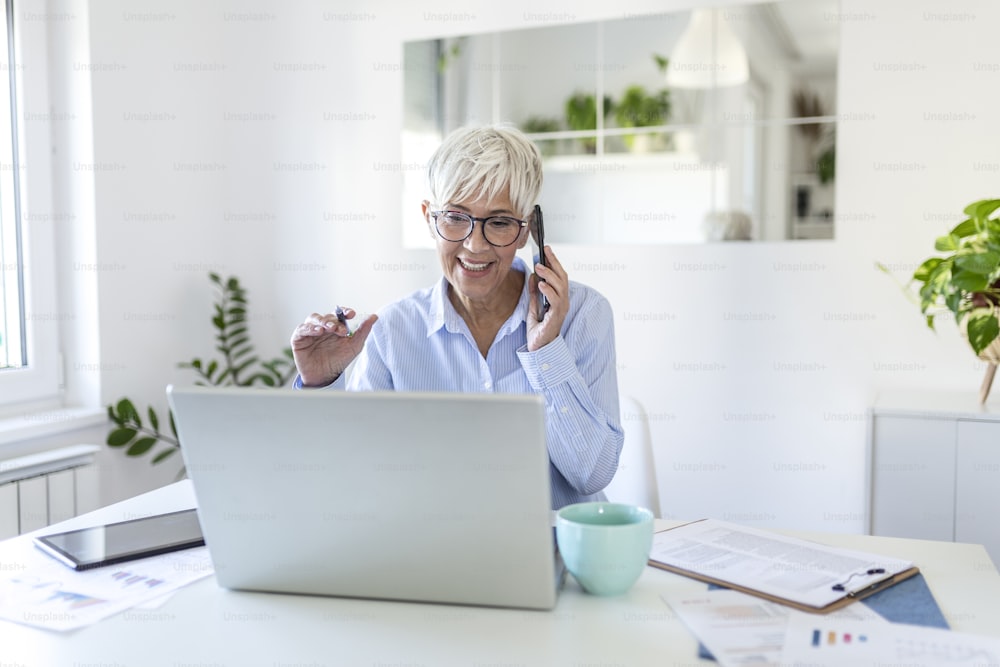 Smiling mature beautiful business woman with white hair working on laptop in bright modern home office. Business woman talking on her mobile phone while working from home