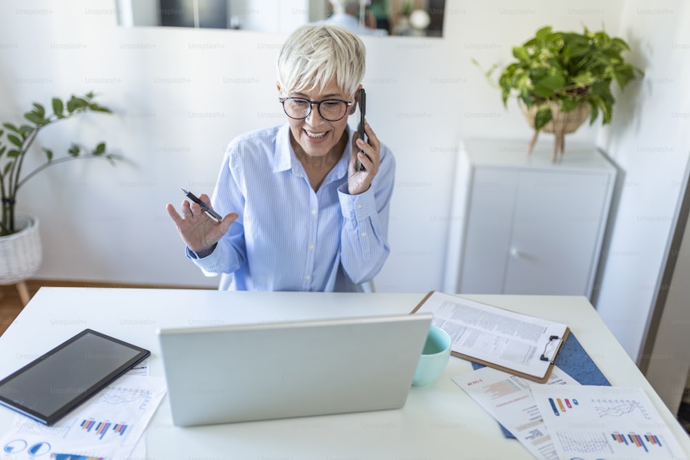 Focused old woman with white hair at home making a business call wile using laptop. Senior stylish entrepreneur wearing eyeglasses working at home. Woman managing domestic bills and home finance